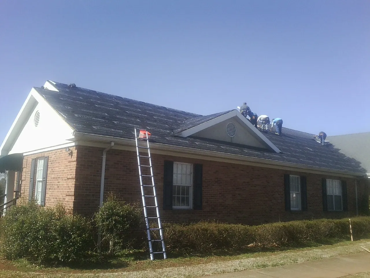 Crew tearing shingles off a brick ranch roof with a ladder set — Metal Roof Repair work by our crew in Newsoms
