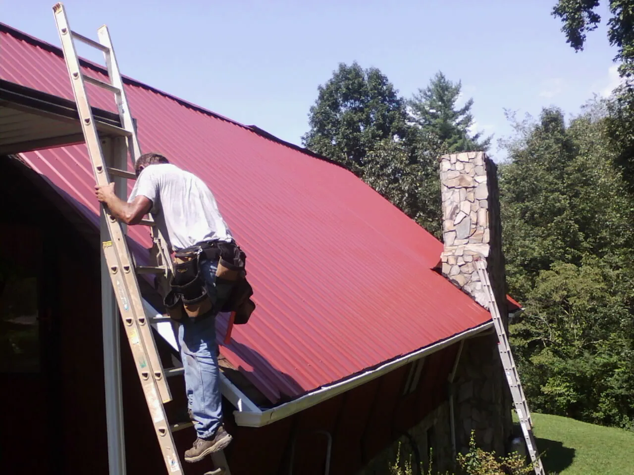 Roofer on a ladder beside a red metal roof and stone chimney — Soffit & Fascia Repair by Roofing Brothers in Newsoms